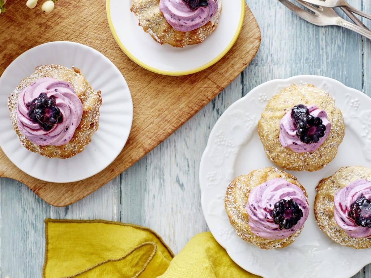 Auf einem Teller stehen 3 Apfelmus-Brombeer-Gugelhupf. Sie sind mit einer lila Creme und je einer Brombeere verziert.