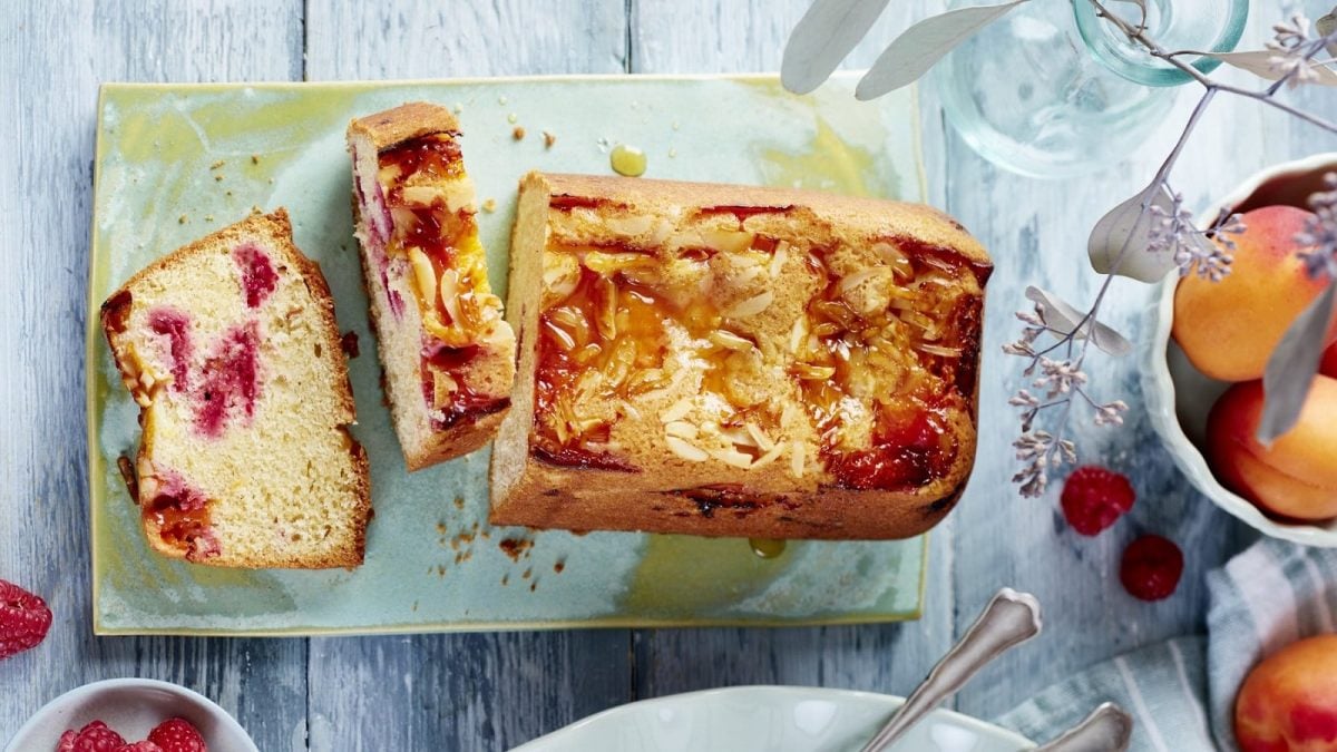 Auf einem Teller steht ein angeschnittener Aprikosen-Himbeer-Kuchen. Er ist mit Mandelsplittern bedeckt.