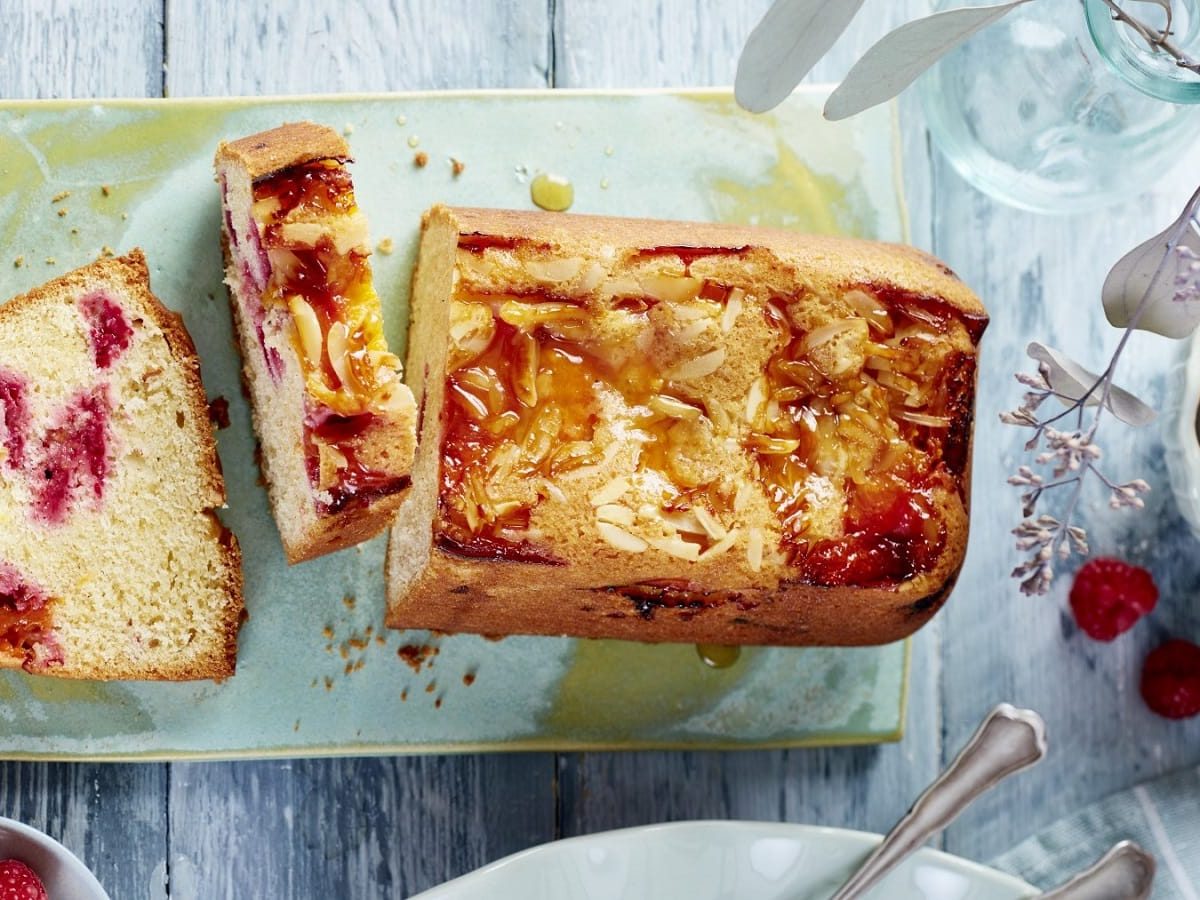 Auf einem Teller steht ein angeschnittener Aprikosen-Himbeer-Kuchen. Er ist mit Mandelsplittern bedeckt.