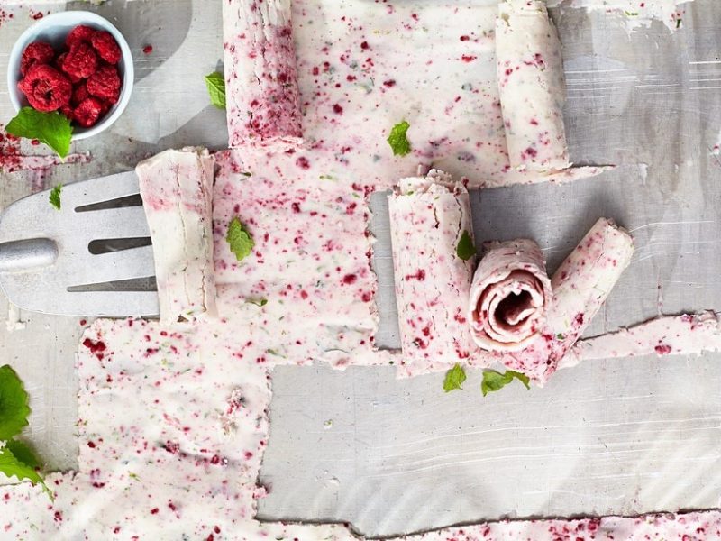 Auf einem Blech ist eine Portion geschabtes Kokos-Limetten-Eis mit Himbeeren ausgestrichen. Es wird mit einem Spatel augerollt.