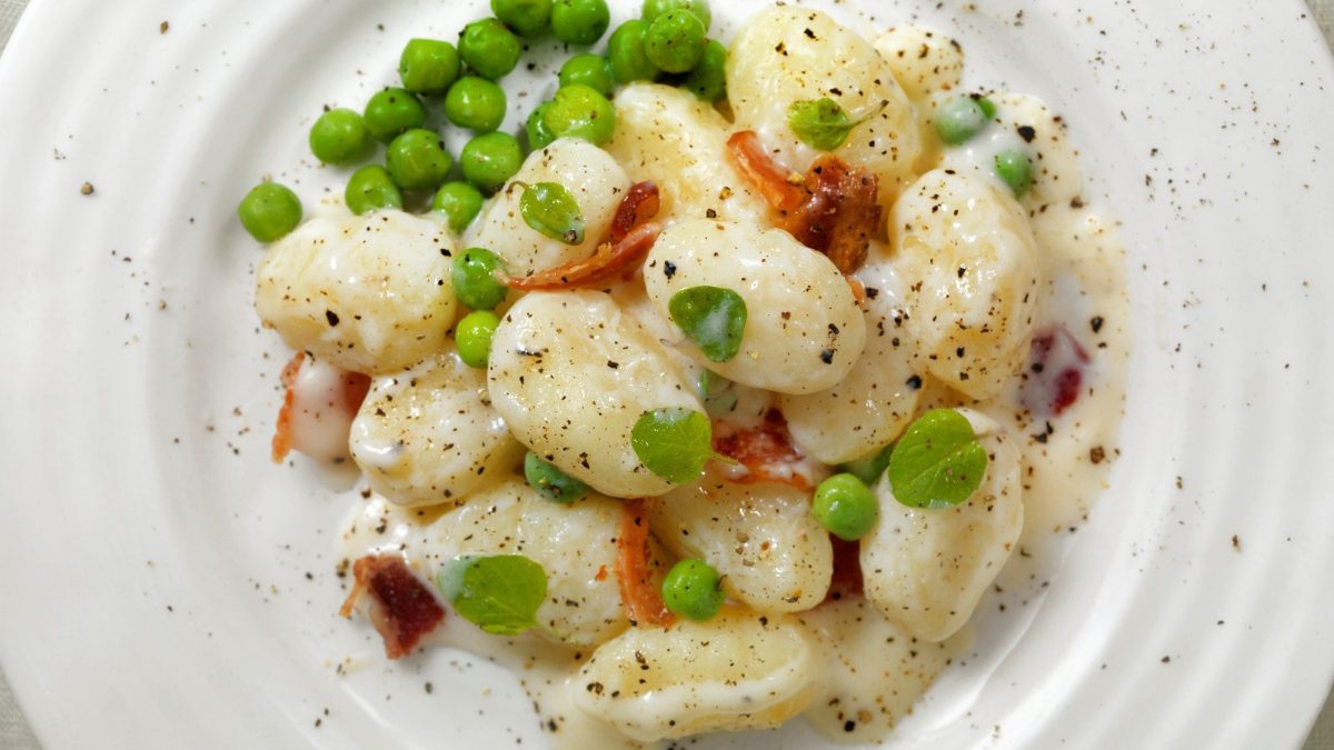 Gnocchi in Knoblauchcreme mit Erbsen und Speck auf weißem teller.