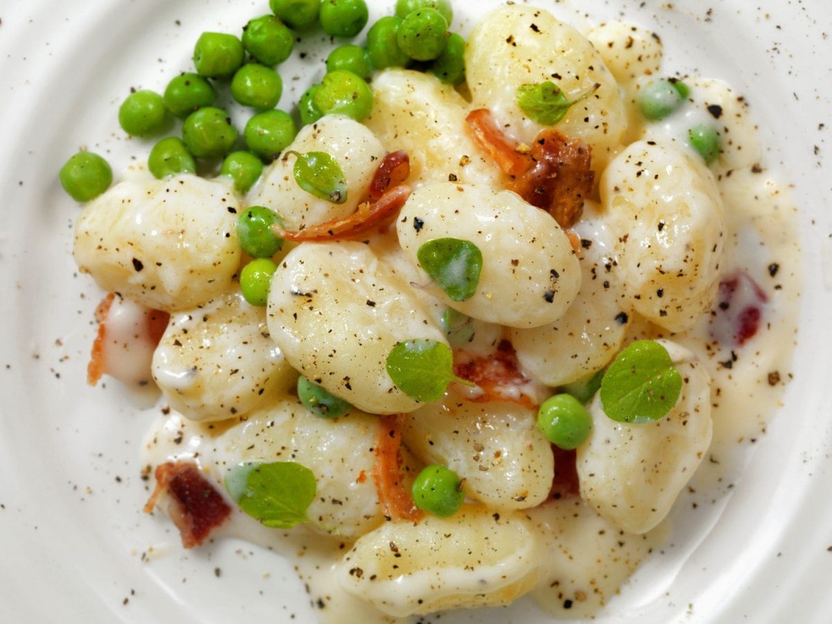 Gnocchi in Knoblauchcreme mit Erbsen und Speck auf weißem teller.