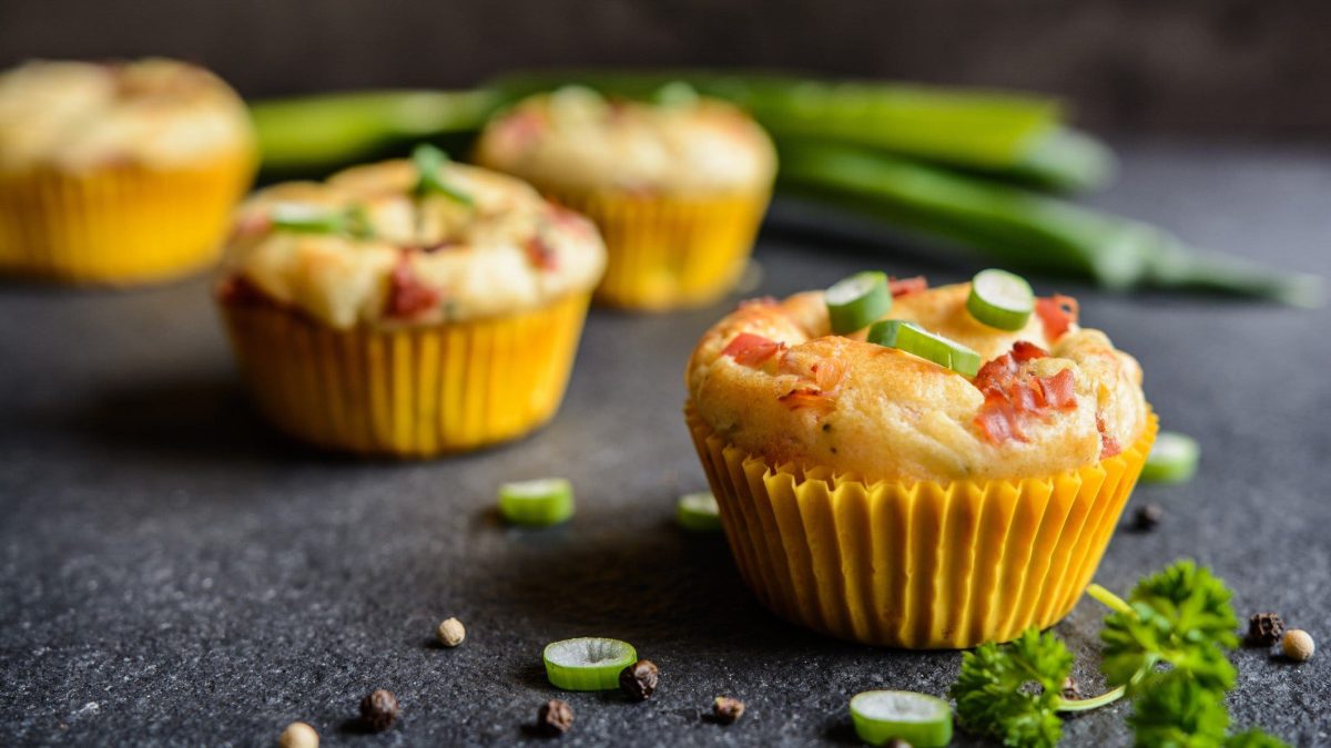 Herzhafte Muffins mit Speck, Käse & Frühlingszwiebeln auf dunklem Untergrund. Daneben liegen Frühlingszwiebeln und Petersilie