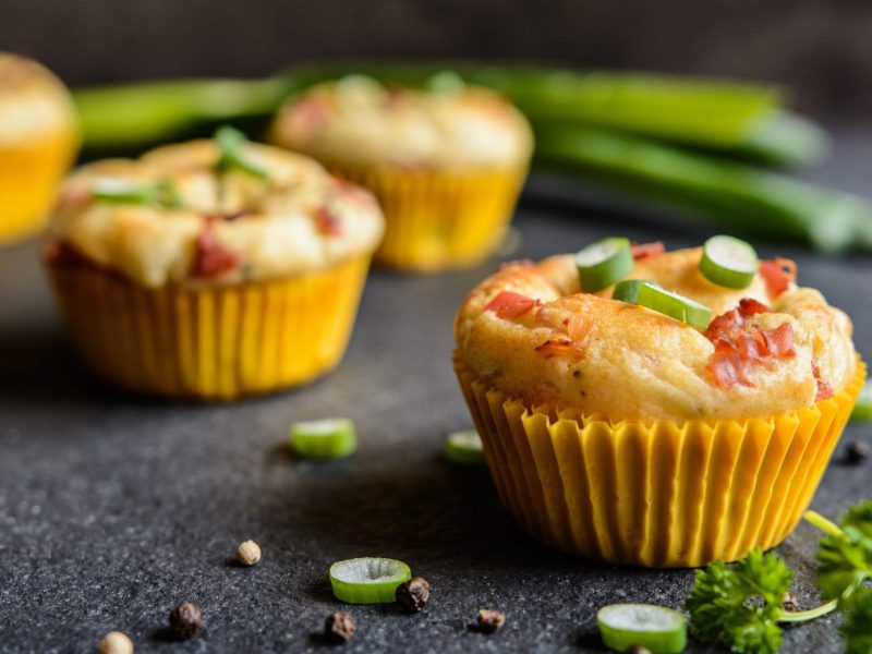 Herzhafte Muffins mit Speck, Käse & Frühlingszwiebeln auf dunklem Untergrund. Daneben liegen Frühlingszwiebeln und Petersilie