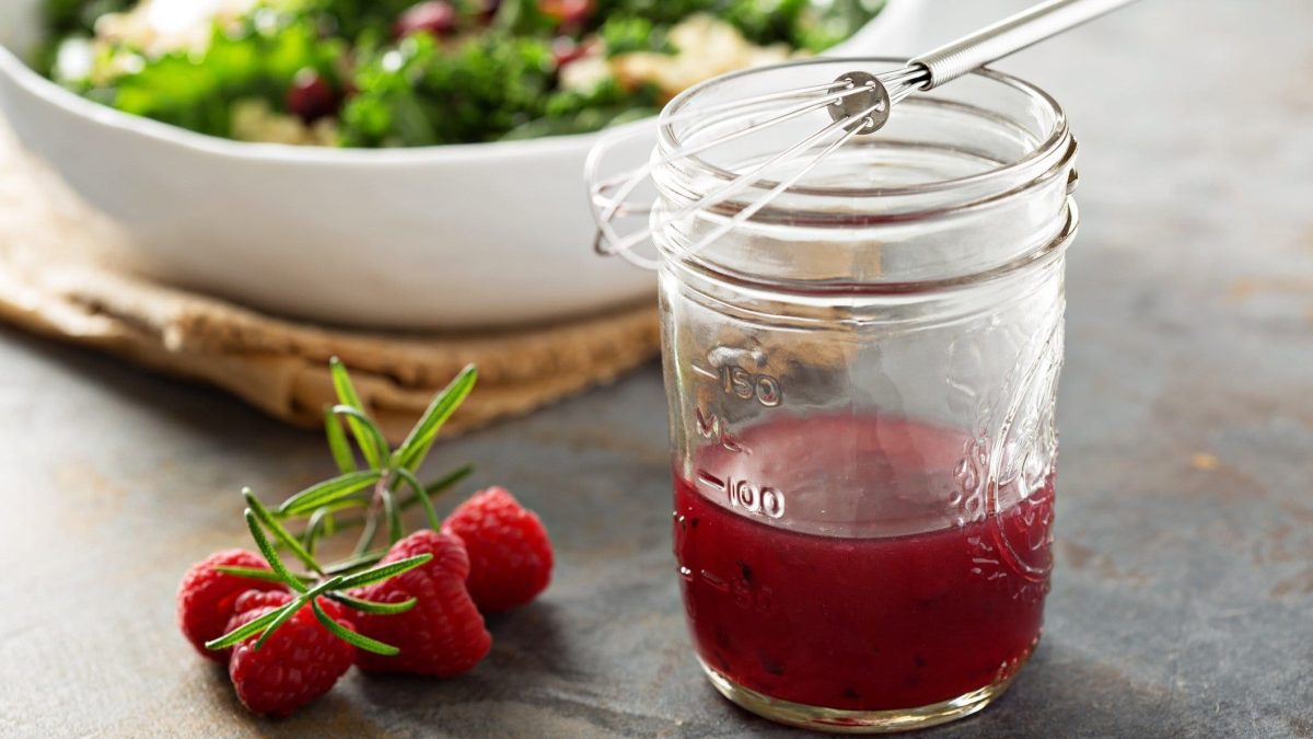 Glas mit Himbeer-Vinaigrette vor einer Schüssel Salat auf dunklem Untergrund.