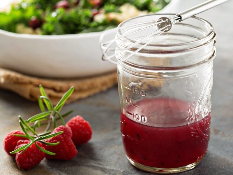 Glas mit Himbeer-Vinaigrette vor einer Schüssel Salat auf dunklem Untergrund.