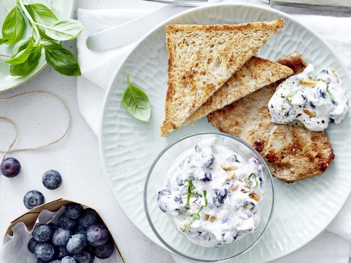 Kalbsschnitzel mit Blaubeer-Dip, bestehend aus Quark, Frischkäse und Pinienkernen, garniert mit frischem Basilikum und gereicht mit getoastetem Toast auf einem hellen Teller. Dazu sind frischen Blaubeeren und Basilikum gereicht.