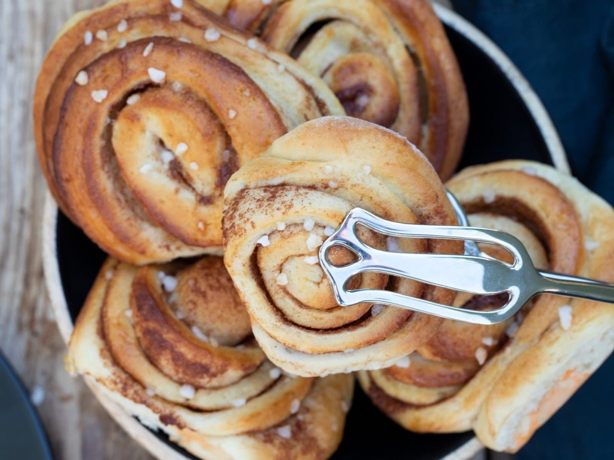 Kanelbullar mit Hagelzucker in einer Schüssel. Eine Zimtschnecke wird mit einer Zange hochgehoben.