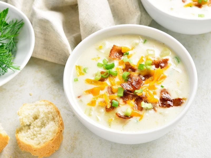 Kartoffelsuppe mit Speck, Käse und Frühlingszwiebeln in weißer Schale auf hellem Untergrund.