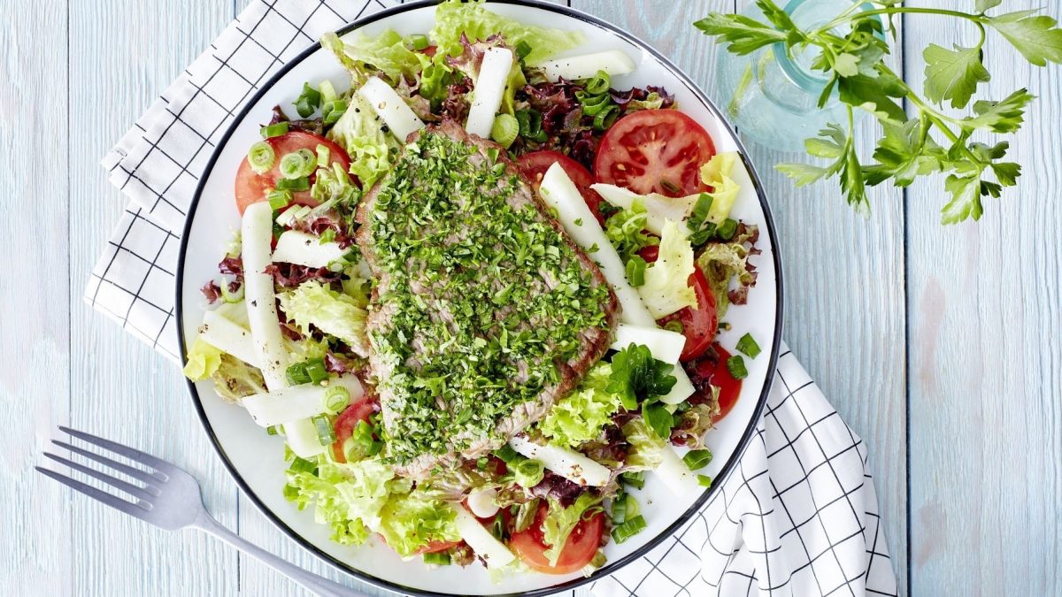 Kräuter-Schnitzel mit Salat mit Tomaten und Kohlrabi, garniert mit frischer Petersilie auf einem hellen Teller auf rustikalem, hellen Holztisch.