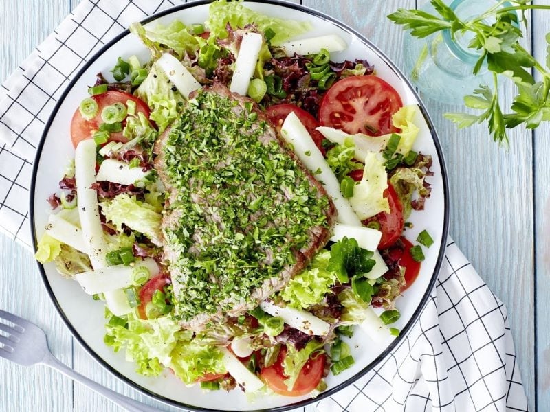 Kräuter-Schnitzel mit Salat mit Tomaten und Kohlrabi, garniert mit frischer Petersilie auf einem hellen Teller auf rustikalem, hellen Holztisch.