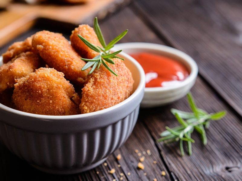 Mini-Schnitzel in weißer Schüssel auf dunklem Holz. Daneben steht ein Schälchen Ketchup.