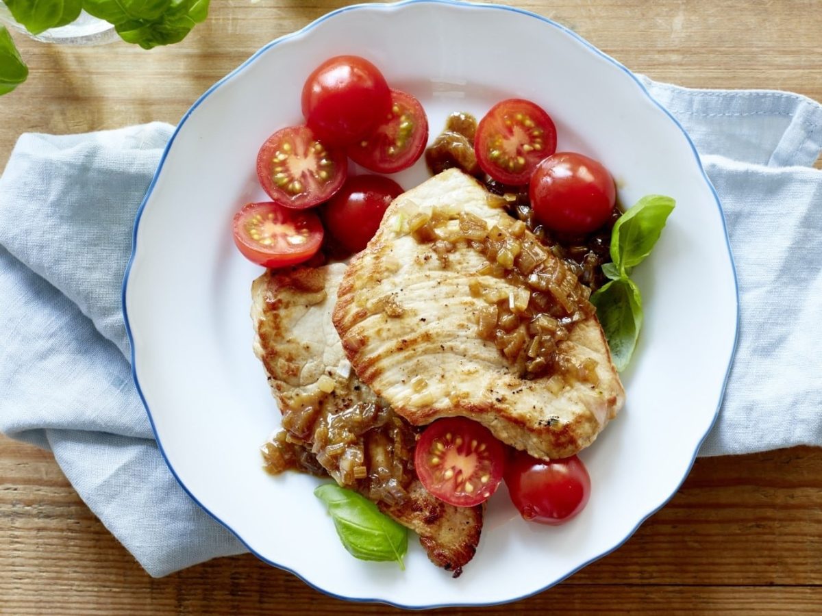 Einfaches Minischnitzel mit Tomate und angerösteten Schalotten, serviert auf einem weißen Teller auf einem Holztisch, garniert mit frischem Basilikum.