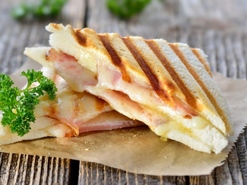 Italienische Panini mit Schinken und Käse auf Brotpapier. Das Ganze liegt auf einem Holz-Untergrund.