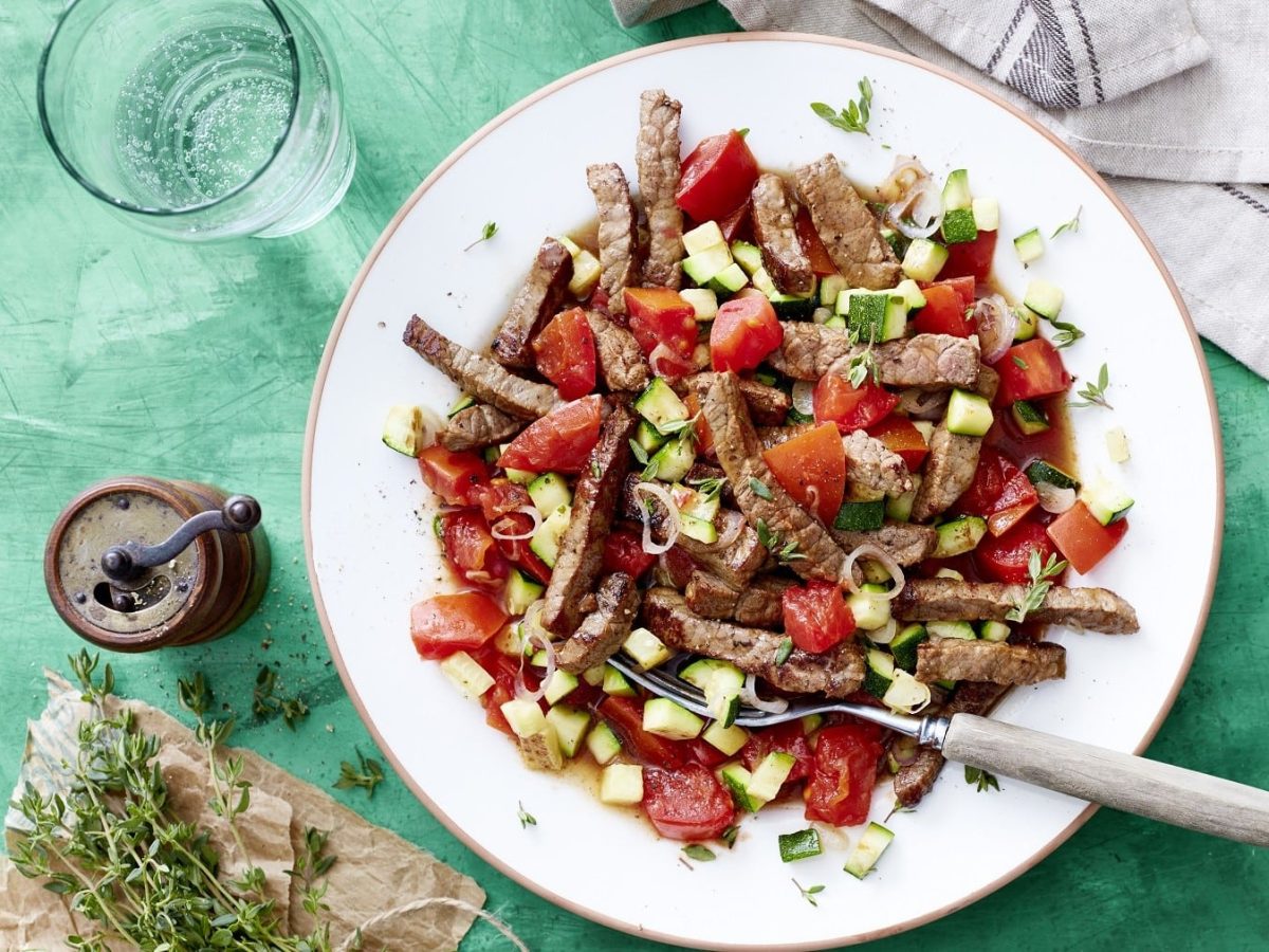 In Streifen geschnittenes Schnitzel mit Tomaten-Zucchini-Sugo und frischem Thymian, serviert in einem weißen Teller auf einem türkisenen Untergrund.