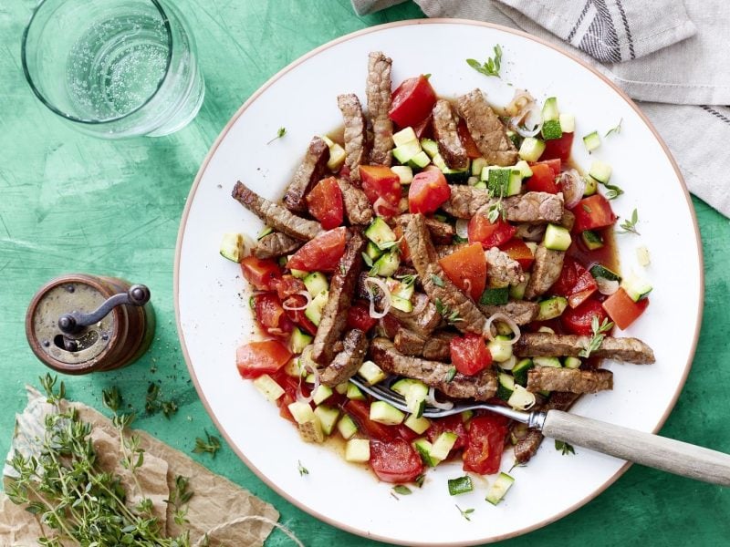 In Streifen geschnittenes Schnitzel mit Tomaten-Zucchini-Sugo und frischem Thymian, serviert in einem weißen Teller auf einem türkisenen Untergrund.