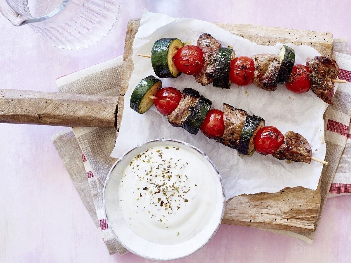 Auf einem Holzbrett liegen Steak-Spieße mit Zucchini und Kirschtomaten. Daneben steht ein Dip.