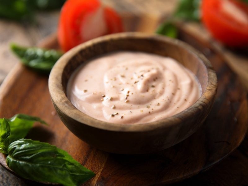 Thousand-Island-Dressing in dunkler Schale auf Holzbrett. Daneben liegen Basilikum und Tomaten.