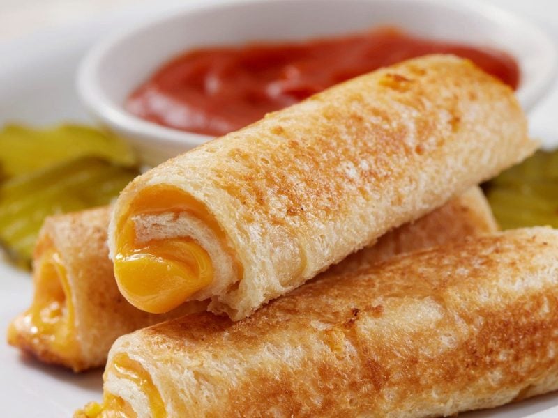 Mehrere gerollte Toast-Brote mit Käsefüllung auf weißem Teller. Daneben ein Tomaten-Dip und Gewürzgurken.