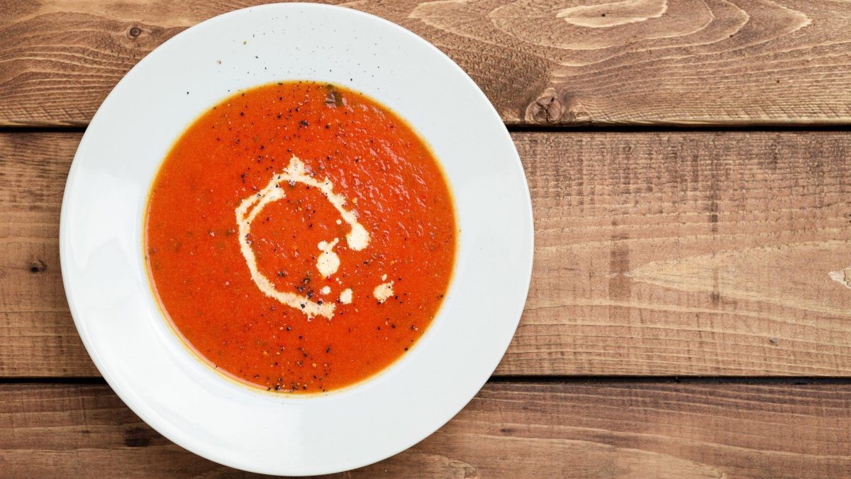 Tomatensuppe mit Kokosmilch und Pfeffer auf weißem Teller, angerichtet auf Holzplatte.