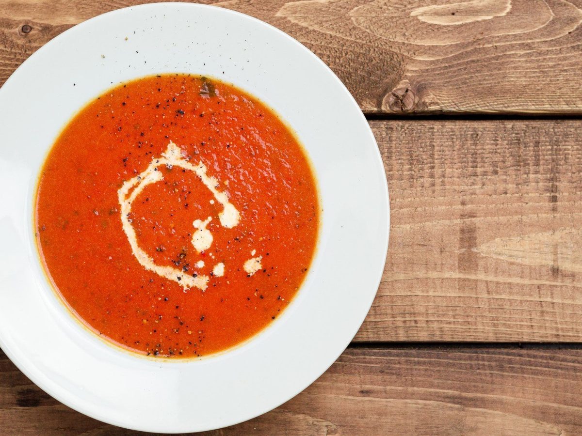 Tomatensuppe mit Kokosmilch und Pfeffer auf weißem Teller, angerichtet auf Holzplatte.