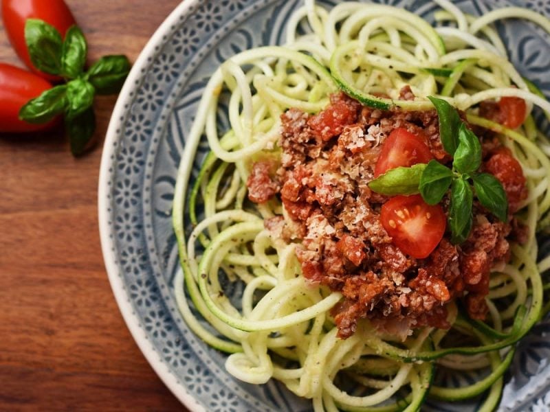 Auf einem Teller liegt eine Portion Zoodles mit Tofu-Bolognese. Sie ist mit vegetarischem Hack und Tomaten garniert.