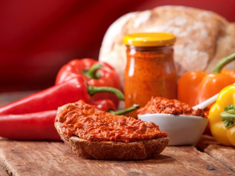 Ajvar auf einer Scheibe Brot vor Gemüse und einem Glas mit Ajvar. Im Hintergrund Brotlaib. Frontalansicht.