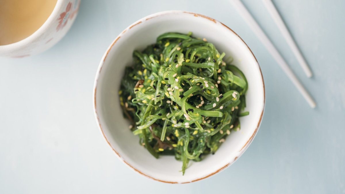 Japanischer Algensalat mit Sesam in einer Schüssel. Daneben eine Tasse Tee und Stäbchen.