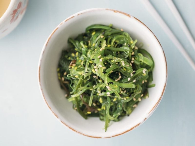 Japanischer Algensalat mit Sesam in einer Schüssel. Daneben eine Tasse Tee und Stäbchen.