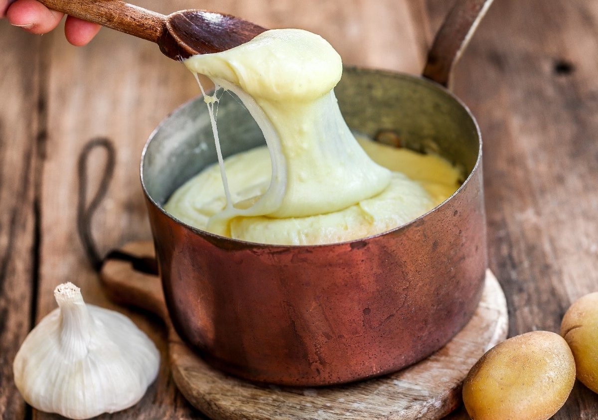 Ein kupferner Topf auf einem Holzbrett mit Knoblauch und Kartoffeln an der Seite. Eine Hand zieht mit einem Kochlöffel etwas von dem Aligot in die Länge.