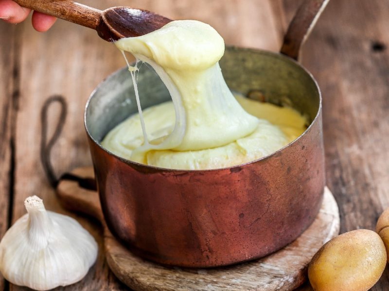 Ein kupferner Topf auf einem Holzbrett mit Knoblauch und Kartoffeln an der Seite. Eine Hand zieht mit einem Kochlöffel etwas von dem Aligot in die Länge.