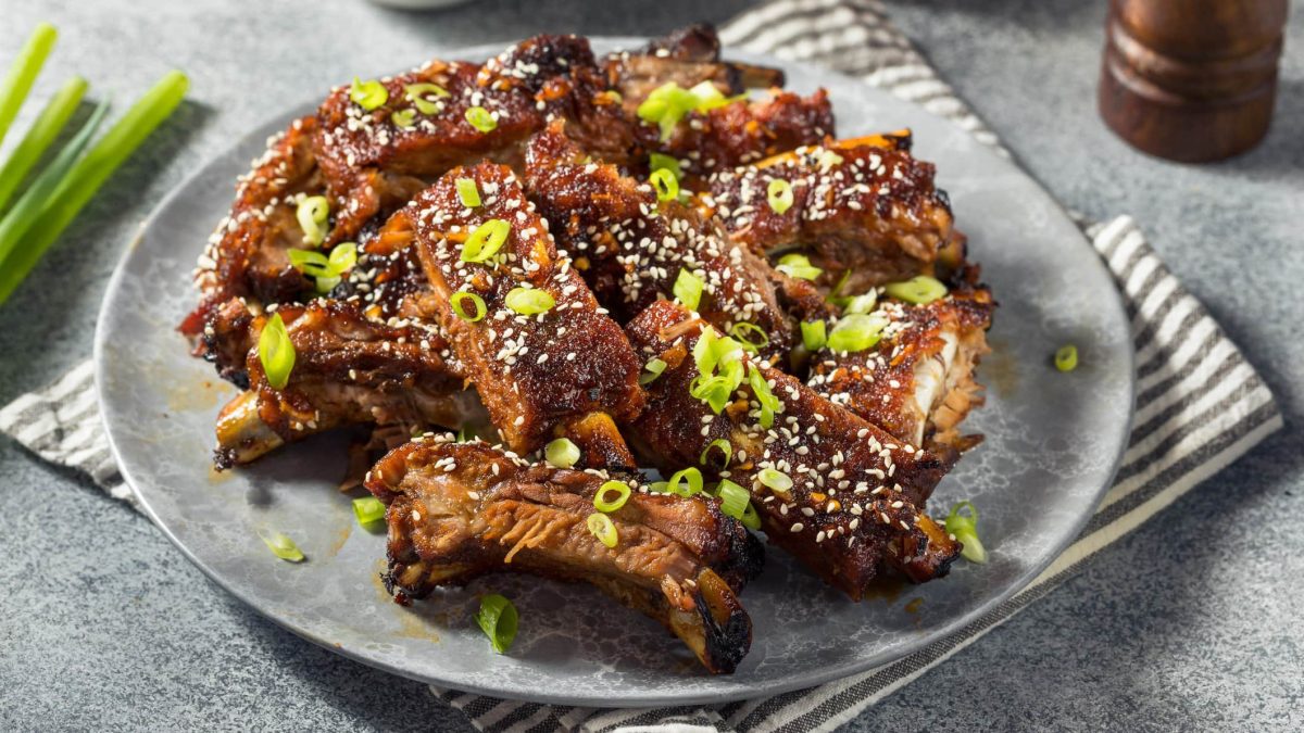 Ein grauer Teller mit den asiatisch marinierten Baby-Back-Ribs auf grauem Untergrund. Auf den Rippchen ist Sesam und Frühlingslauch.