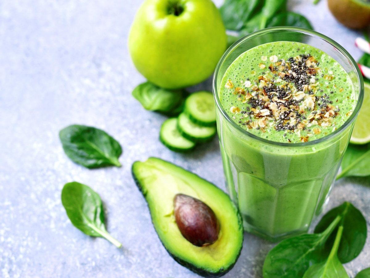 Ein grünes Glas mit dem Smoothie auf grauem Untergrund. Drum herum viel grünes Obst und Gemüse.