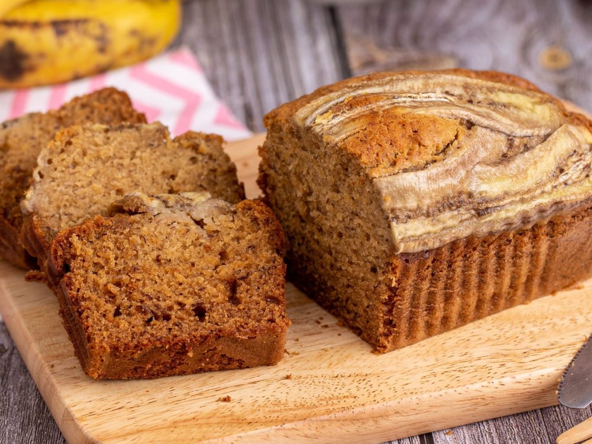 Selbst gemachtes Bananenbrot auf Holzbrett und grauem Holztisch. Oben reife Bananen und rot-weißes Stofftuch. Unten rechts Messer. Drei Scheiben geschnitten auf dem Brett trappiert. Frontalansicht.