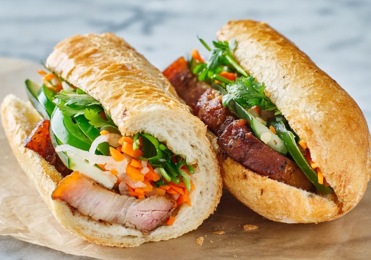 Zwei Banh-mi Sandwiches auf einem Marmortisch mit etwas Backpapier. Das Sandwich ist angeschnitten, sodass man den Inhalt (Karotten, Paprika, Schweinefleisch und Koriander) sehen kann.