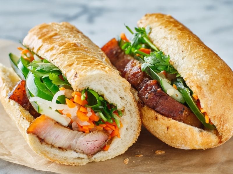 Zwei Banh-mi Sandwiches auf einem Marmortisch mit etwas Backpapier. Das Sandwich ist angeschnitten, sodass man den Inhalt (Karotten, Paprika, Schweinefleisch und Koriander) sehen kann.