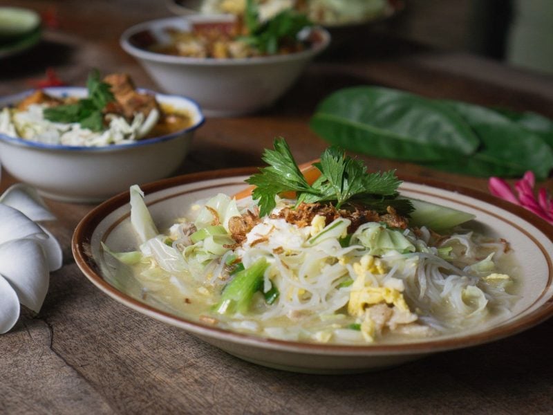 Bihunsuppe Bihun Sup Ayam in Suppenteller auf Holztisch. Blüten und Schalen mit Deko im Hintergrund.