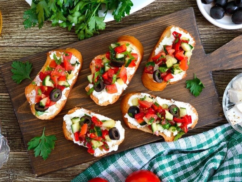 Auf einem Holzbrett liegen Bruschetta mit Ziegenkäse und frischem Gemüse.