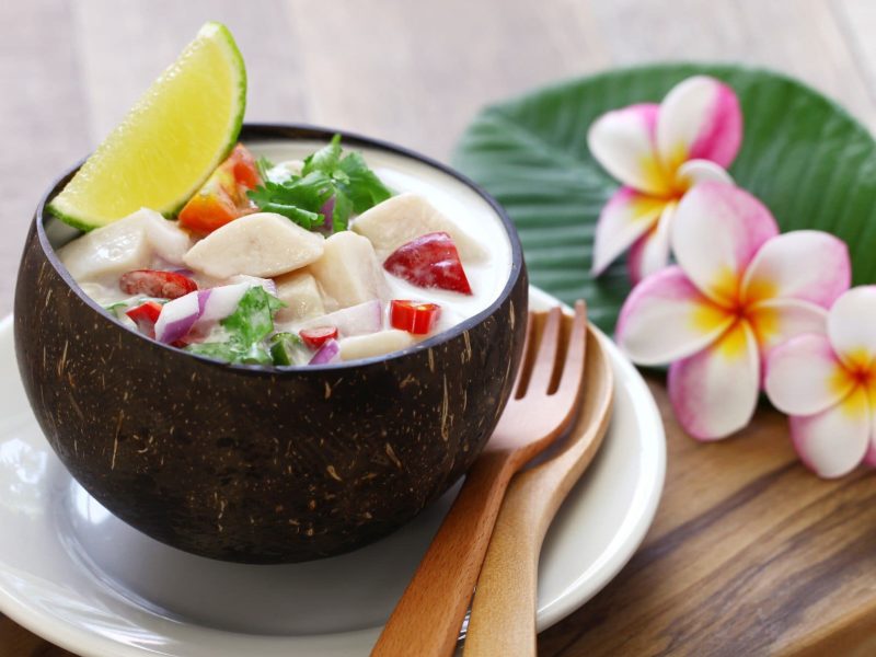 Exotische Ceviche mit Kokosmilch und Chili in Kokosschale auf weißen´m Teller auf Holzbrett. Daneben Holzbesteck und Blütendeko. Draufsicht.