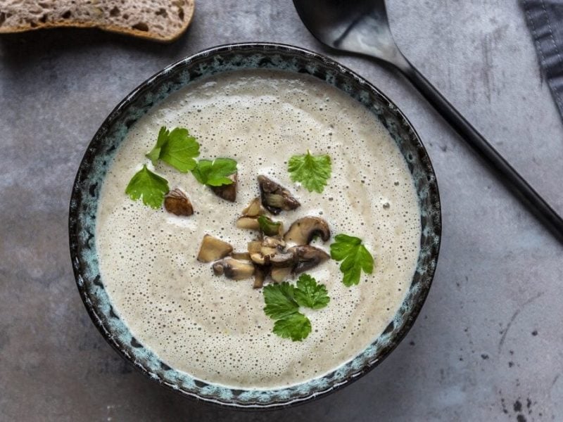 Eine Teller Champignon-Erdnuss-Suppe steht auf einem Holztisch. Daneben liegen ein Suppenlöffel und eine Scheibe Brot.