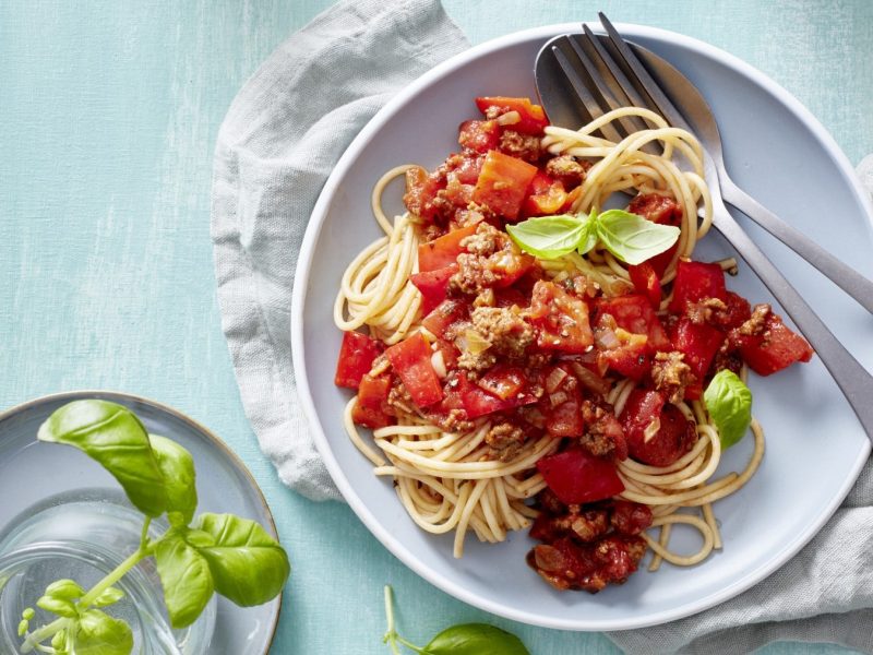 Chili-Pasta mit Hack und Paprika auf einem weißen Teller. Daneben Besteck und Basilikum.