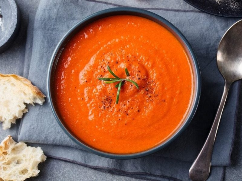 Eine Schüssel mit einer Curry-Gazpacho steht auf einem Tisch. Daneben liegen Weißbrotscheiben und ein Löffel.