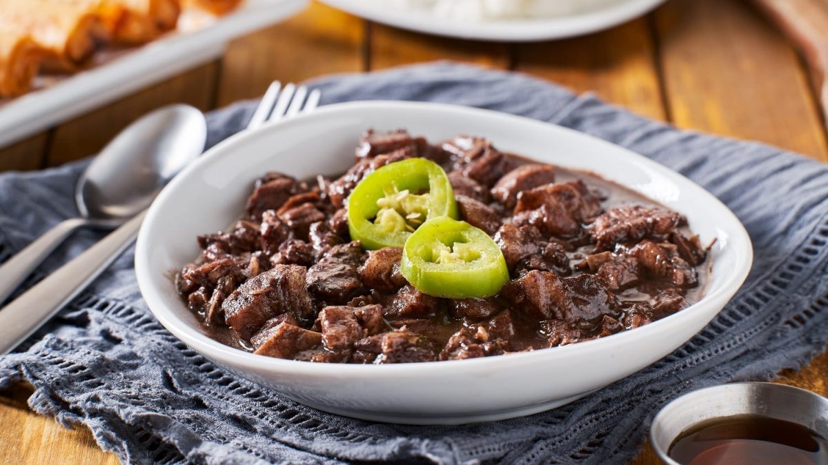 Eine weiße Schale mit Dinuguan und grüner Peperoni auf einem Holztisch mit grauem Tuch. Oben links im Bild ist Besteck und ein weiterer Teller.