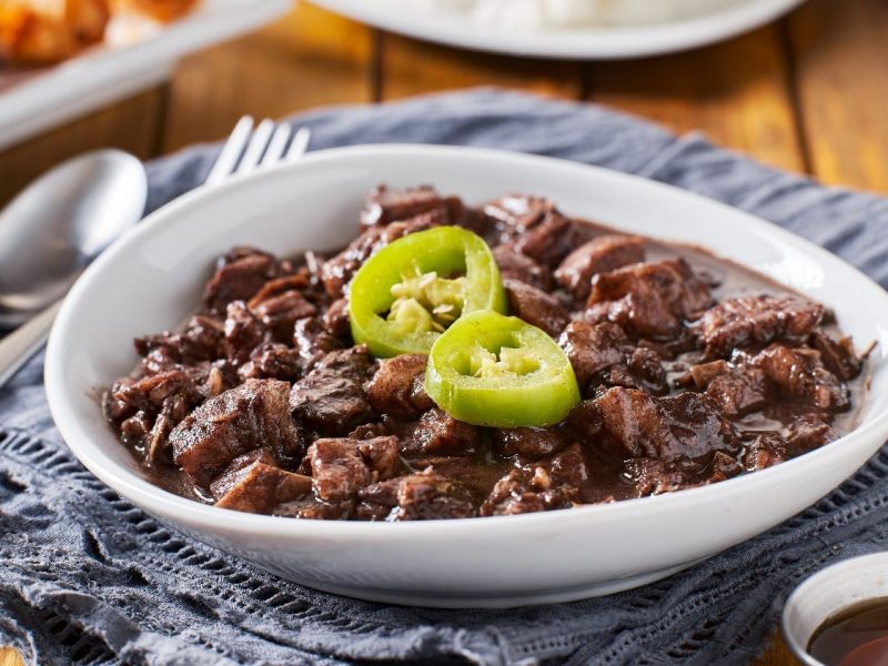 Eine weiße Schale mit Dinuguan und grüner Peperoni auf einem Holztisch mit grauem Tuch. Oben links im Bild ist Besteck und ein weiterer Teller.