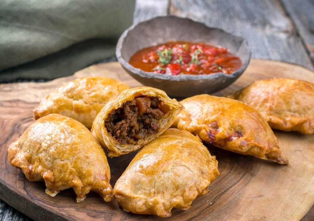 Fünf Empanadas auf einem Holzbrett und grauem Hintergrund. Hinter dem Brett ist eine Dip-Schale mit einer roten Salsa.