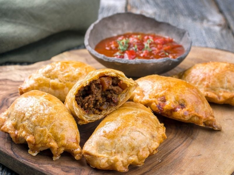 Fünf Empanadas auf einem Holzbrett und grauem Hintergrund. Hinter dem Brett ist eine Dip-Schale mit einer roten Salsa.