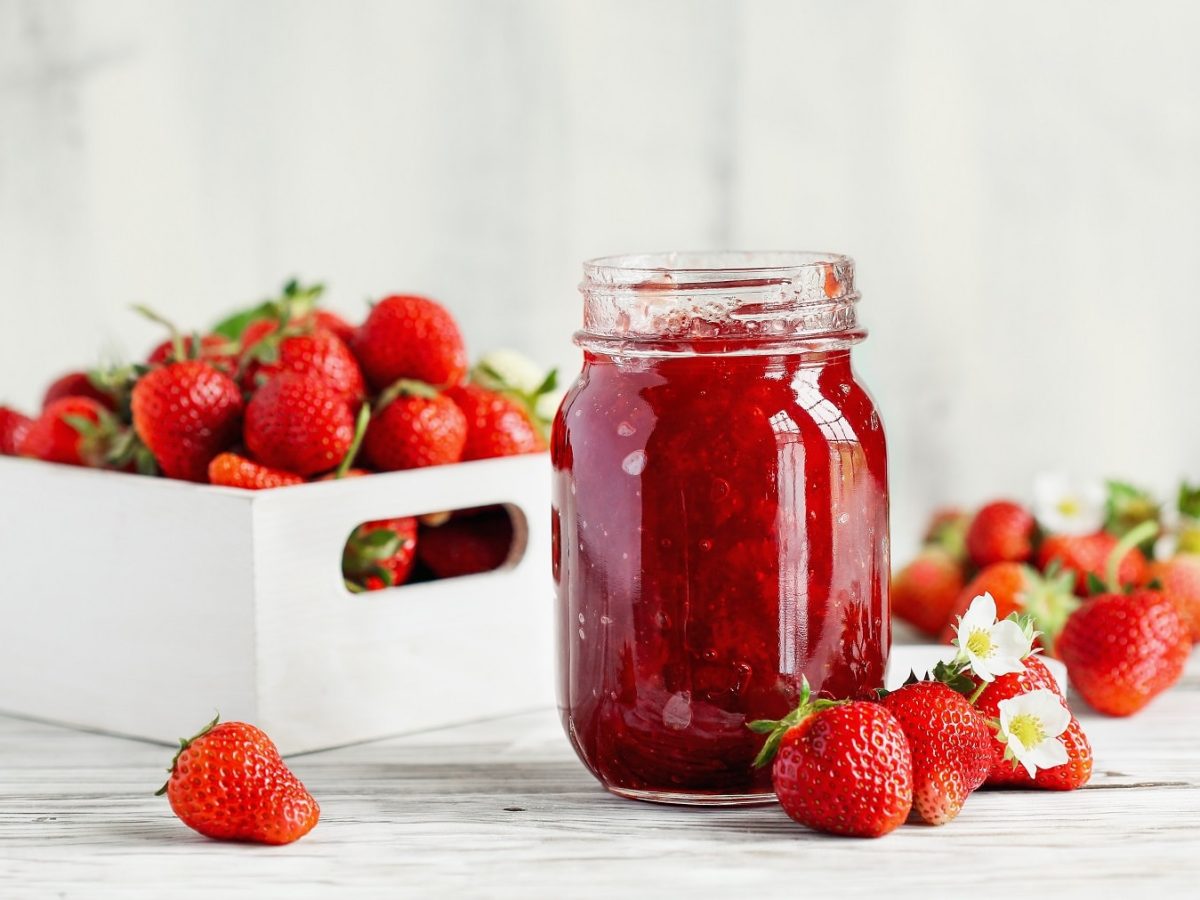 ein Glas Erdbeer-Kokos-Marmelade, dahinter eine weiße Holzkiste mit frischen Erdbeeren.