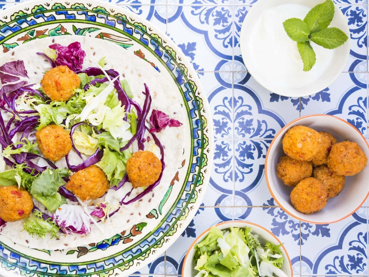 Ein Teller mit Falafel, Salat und Hummus steht auf einem arabisch verzierten Untergrund. Daneben stehen eine SChale mit Falafel, sowie eine Schale mit Salat.