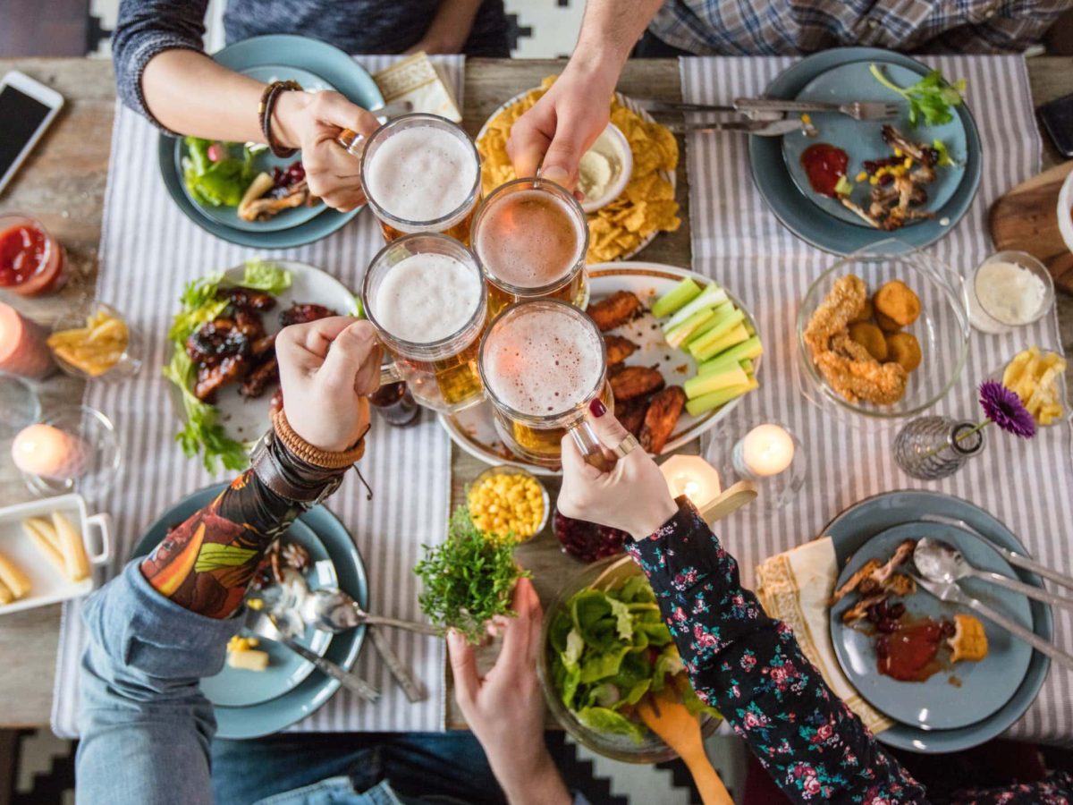 Wie viel Fleisch pro Person soll man kaufen? Freunde stoßen über gedecktem Tisch mit diversem Speisen.