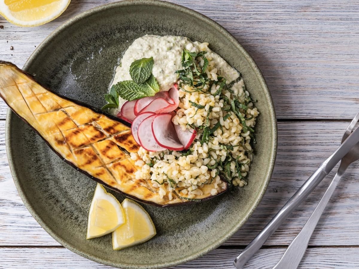 Ein Teller gebackene Augerbine mit Bulgur-Spinat-Salat, Radieschen, Minze und Zitronenspalten in der Draufsicht, daneben Besteck.